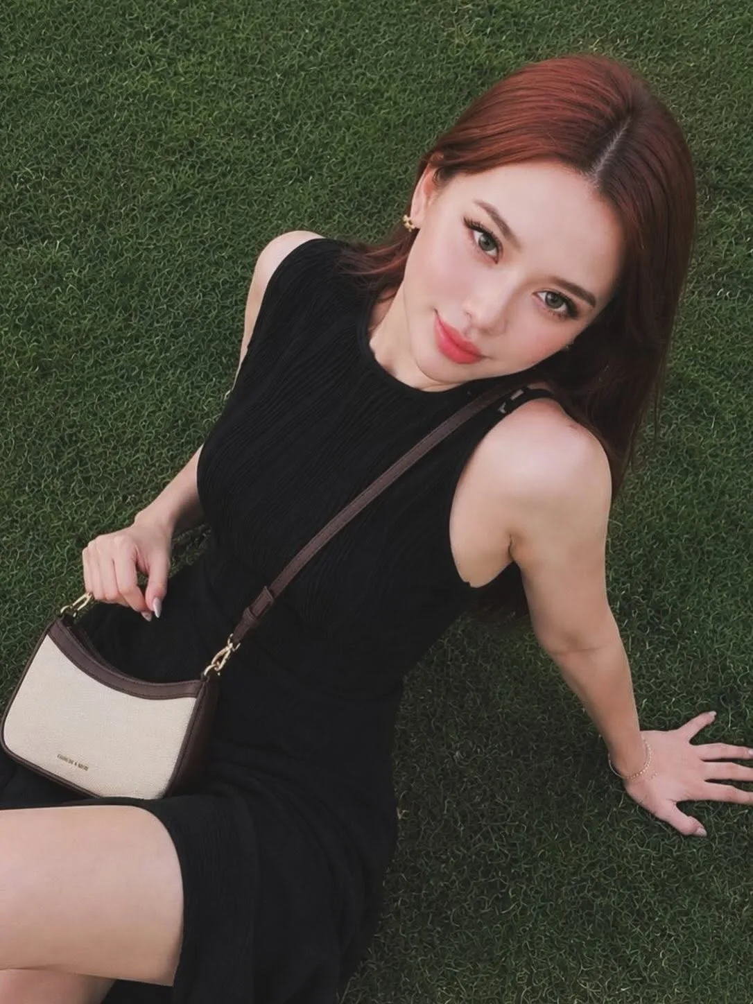 A woman with long auburn hair, glossy and freshly styled, sitting on grass in a black dress.